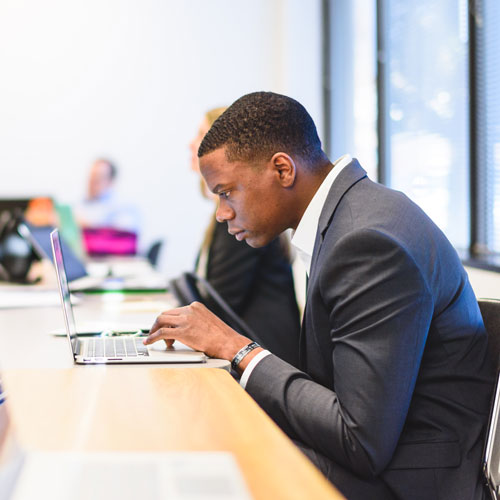 Business man sits in class in a suit, hunched over his laptop.