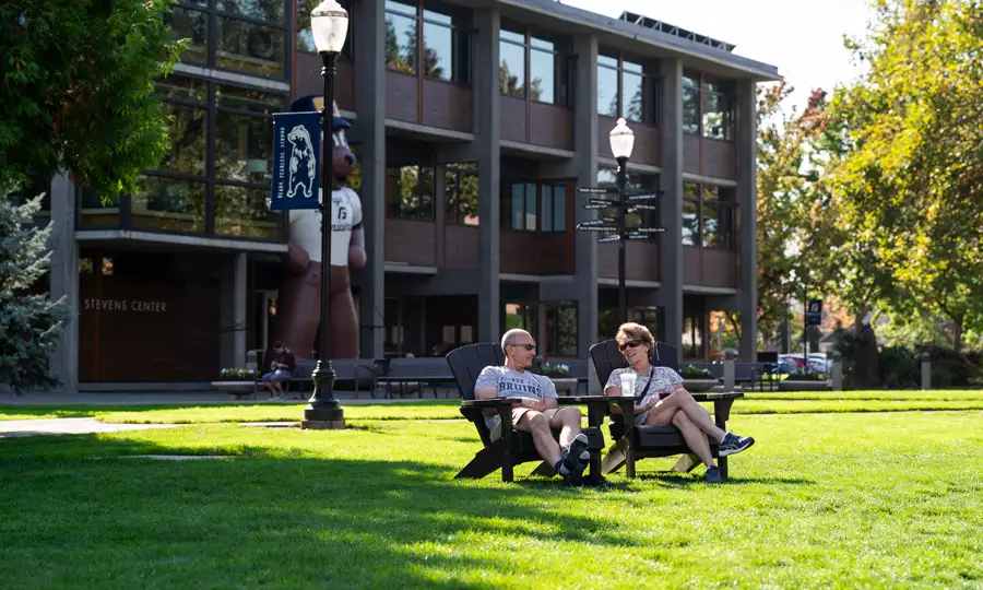 Two alumni sitting on lawn chairs in the sun talking in front of Stevens