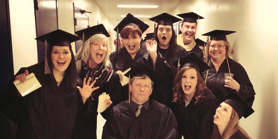 In their graduation attire, Rachel White's Adult Degree Program cohort gathers in a hallway for a photo.