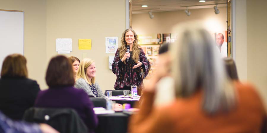 Centered in the image is Dr. Jennie Harrop, who is standing in a table-filled room. She is holding a microphone to her mouth and smiling. Seated at the tables around her are smiling adults.