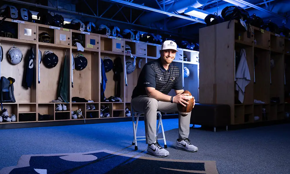 Coach Crace sits in the football locker room, smiling with a football