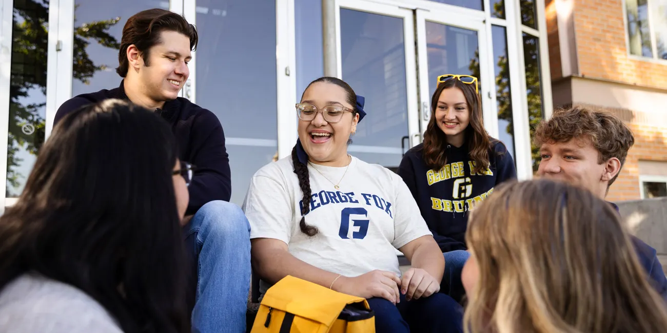 6 students talk and laugh on the steps in front of a building on campus.