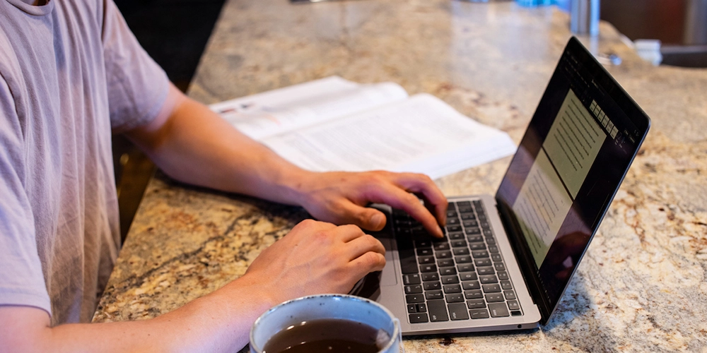 A person using a laptop at a counter with an open book and a cup of coffee nearby.