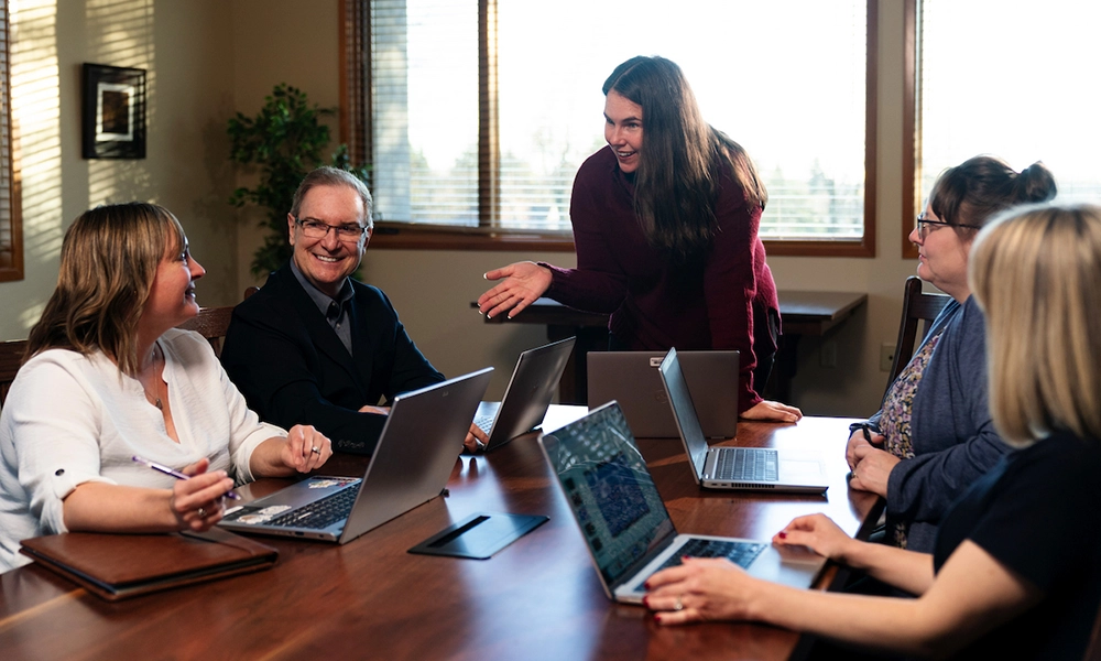 A group of coworkers gathering around a conference table with laptops, discussing and collaborating during a meeting.