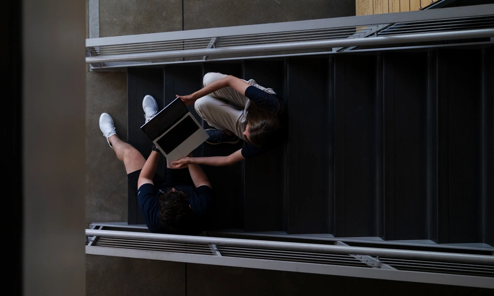 Overhead view of one student passing their laptop to another student while sitting in a stairwell