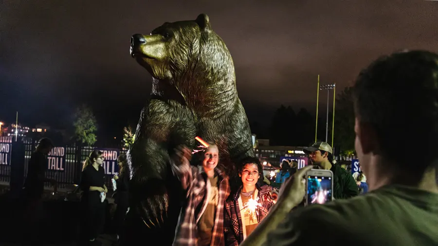 Two George Fox students pose for a photo with sparklers at night with Mama Bear