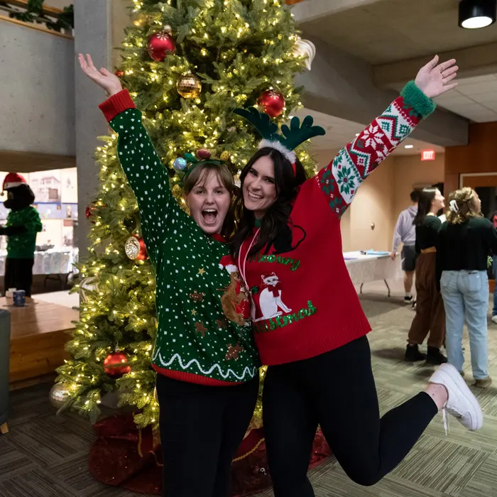 Two student pose in Christmas sweaters in front of a tree