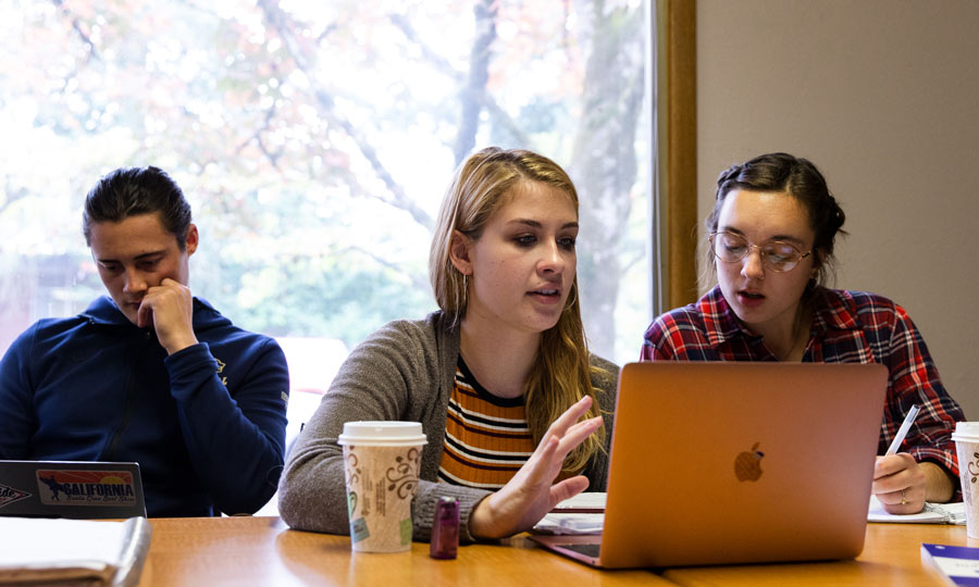 A student at a table with other students talks with someone out of frame with an expression of interest