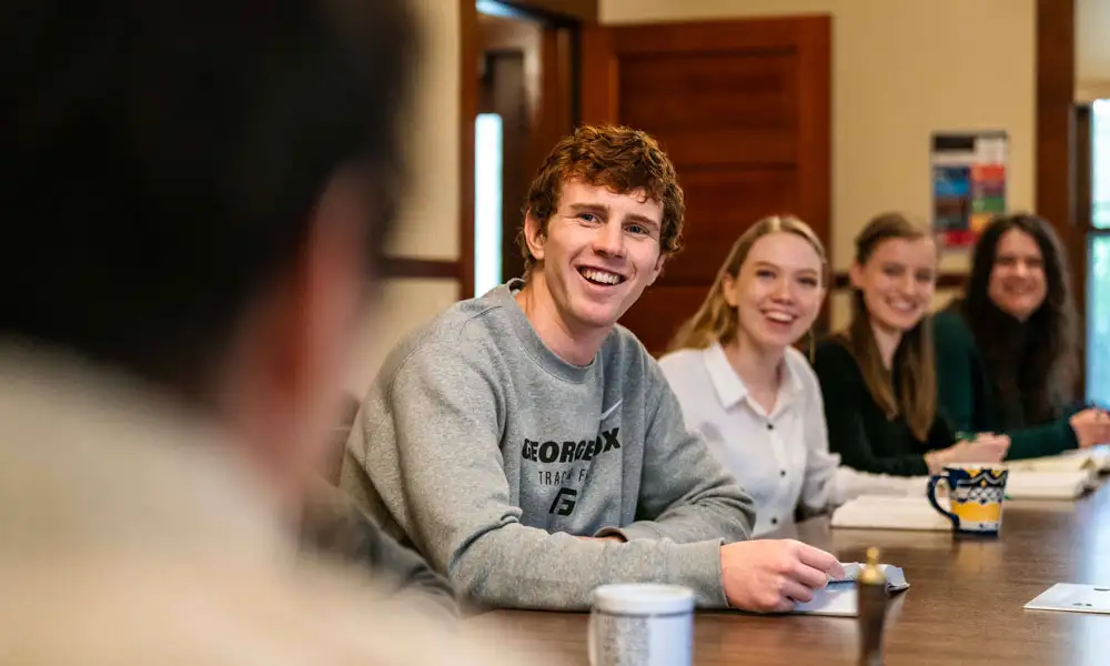 A line of students sitting at a big table with books and mugs in front of them smiling in discussion