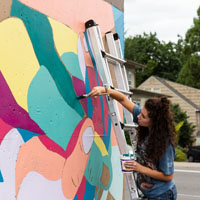 Student standing on a ladder painting a mural on a building wall