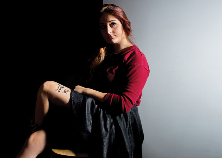 Photo of Bekah Miles sitting in a chair looking at the camera showing the tatoo on her leg. Half of her face is covered in shadow.