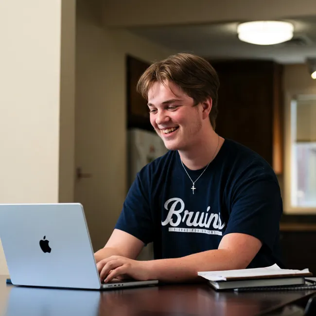 Student with a warm smile works on his laptop at the dining table