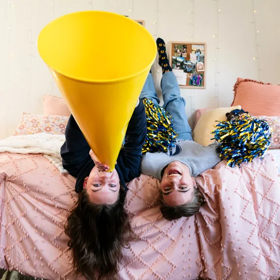 Two roommates lie in a bed upside down and shout through a yellow megaphone