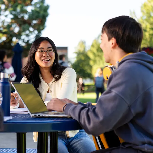 Two students chat and work at a picnic table outside