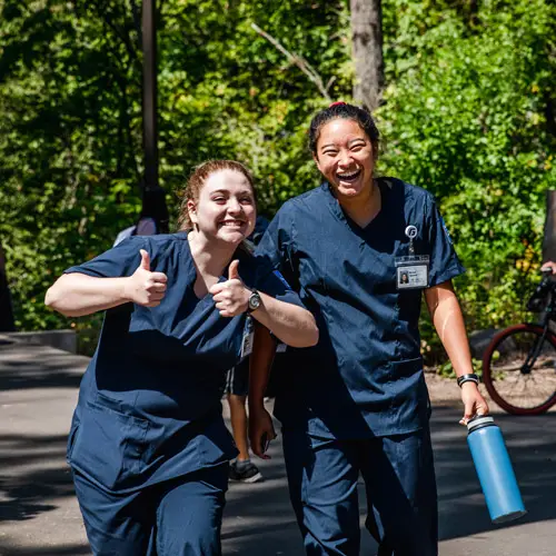 Two nursing students in scrubs smile and wave at the camera