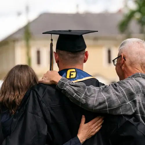A behind shot of a student wearing his George Fox grad cap and gown with his arms around his parents