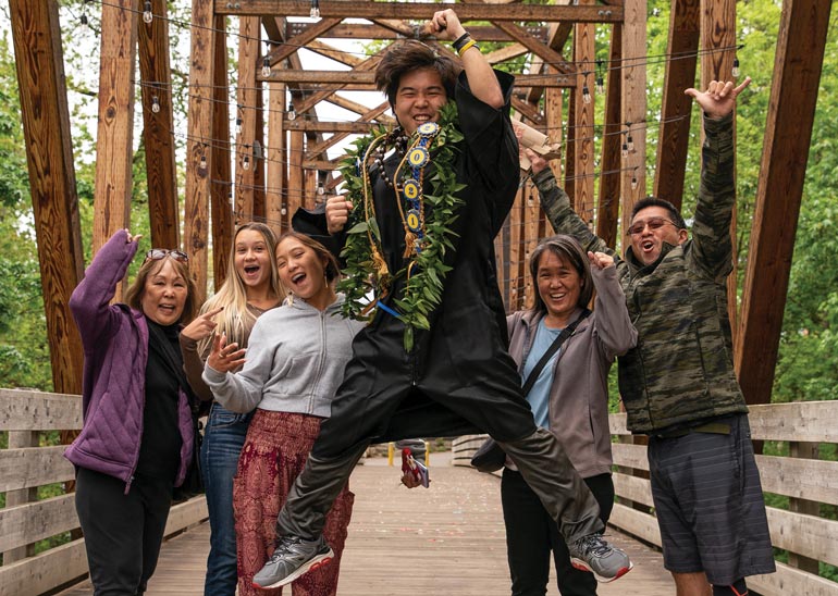 A family stands on the bridge, while a student wearing graduation robes and a hawaian wreath jumps up in celebration in the center