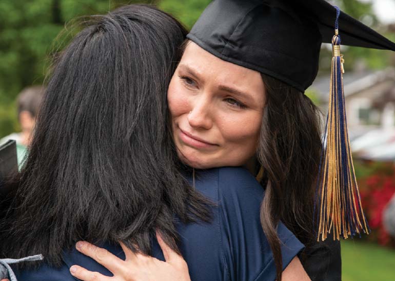 A student in graduation robes holds back tears while she hugs someone