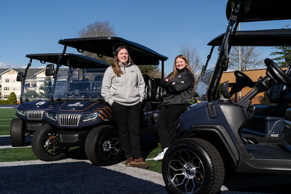 Demi Guild and Jensyn Lown leaning up against the gold carts on the football field