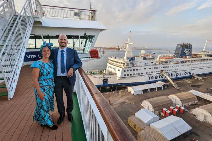 Joff Williams and his wife stand on a deck with a Mercy Ship in the background