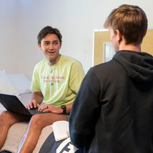 Student working on his laptop on his bed and chatting with his roommate