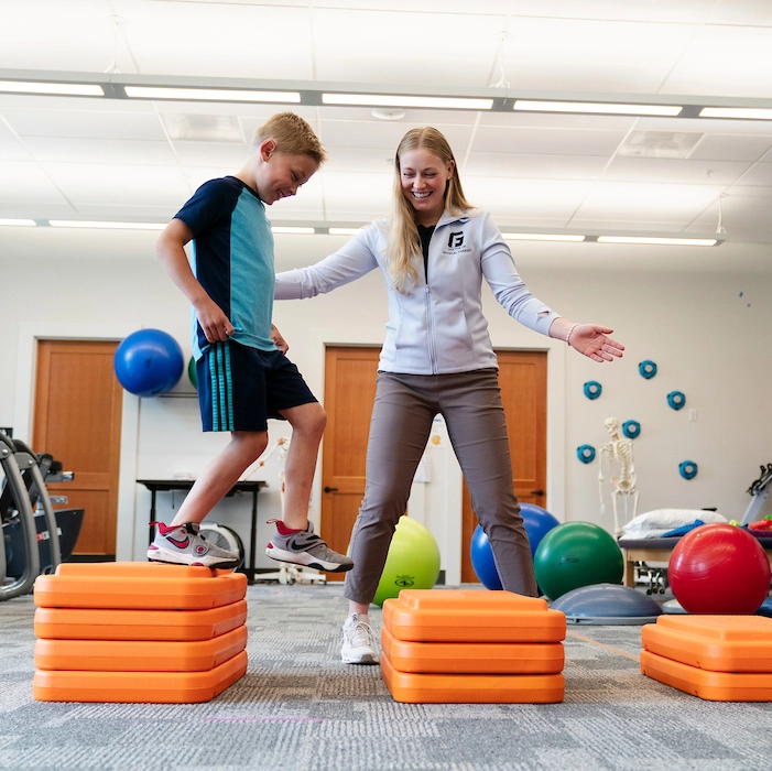 DPT student stands closely beside a kid with her arms outstretched, ready to assist him as the kid jumps along stacked orange exercise steps