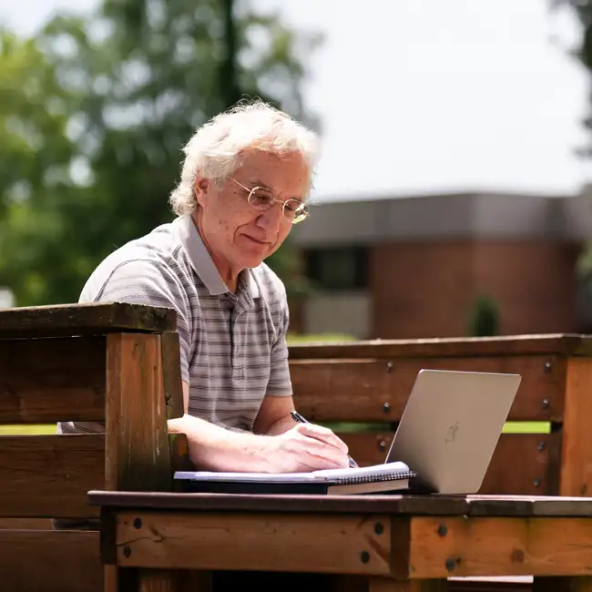 Man writing and working on his laptop in a beautiful outdoor setting on a wood bench